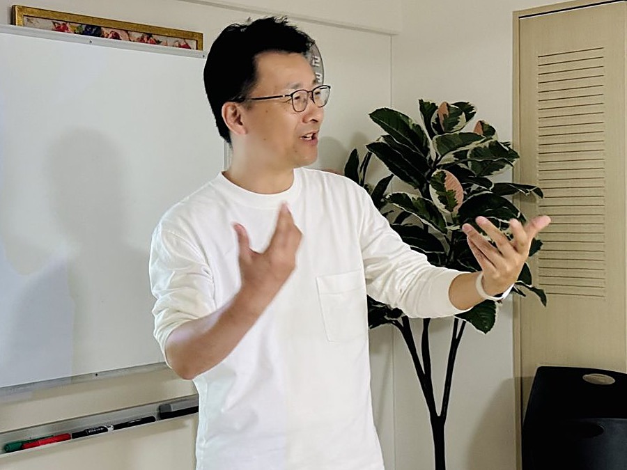 ホワイトボードの前で、手を動かしながら話す伊藤ゆきひろ。白いシャツ姿で、後ろには観葉植物がある